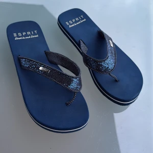 Blå sandaler från Esprit med glitter - Snygga blå sandaler från Esprit med glittrande remmar och diskret mönster. Mjuka och bekväma med platt sula och märkets logga på remmen. Perfekta för varma dagar och chill vibes.