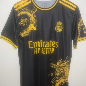 Real Madrid svart/gul/guld fotbollströja M - Snygg svart Real Madrid fotbollströja från Adidas med gula detaljer och drakmotiv. Tröjan har korta ärmar, gula ränder på axlarna och tryck med nummer 7 på ryggen. Materialet är lätt och andas, perfekt för match eller träning.