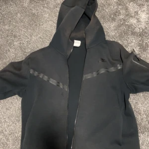 Svart hoodie med dragkedja och huva - Svart hoodie med hel dragkedja och huva. Snygga svarta detaljer i form av band över bröstet och på ena ärmen. Perfekt för en chill och sportig look. Långärmad och med praktisk ficka på ärmen.