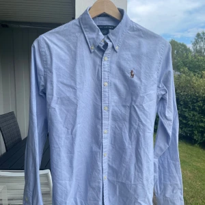 Ljusblå skjorta Ralph Lauren - Klassisk ljusblå skjorta från Ralph Lauren med broderad logga på bröstet. Skjortan har button-down krage, vita knappar och lång ärm. Tillverkad i bomull. Inga fläckar eller skador. Passar normalt, säljer pga för liten storlek. Storlek 8 vilket kan motsvara XS