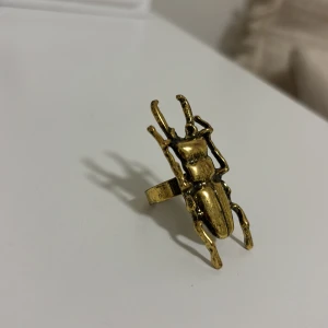 Statementring i guld med insektmotiv - Cool ring i guldtonad metall med en stor insekt som motiv. Ringen har en unik och edgy design som verkligen sticker ut och passar perfekt för dig som gillar att göra ett statement med dina accessoarer.