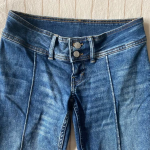 Blå lowwaist bootcut jeans - Snygga blå jeans med bootcut-modell och markerade pressveck längs benen. Jeansen har dubbla knappar i midjan och klassiska sömmar i kontrastfärg. Perfekta för dig som gillar en retrovibe och vill ha ett par jeans som sticker ut lite extra. Hör av dig för frågor. Midjemått rakt över: 37cm men stretchiga. Innerbenslängd: 76cm.🥰