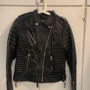 Svart skinnjacka från Boda Skins - Riktigt snygg svart skinnjacka från Boda Skins med cool quiltad design på axlar och nederkant. Jackan har flera dragkedjor, bältesdetaljer och långärmade ärmar med ribbade partier. Perfekt för dig som vill ha en edgy och stilren look.