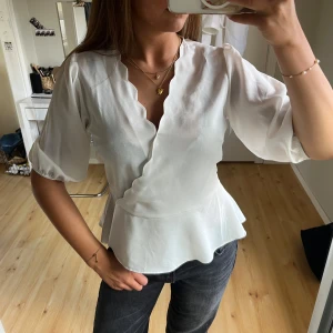 Vit omlottblus med volangkant - Supersöt vit omlottblus med scallop-detaljer längs v-ringningen och volangkant nedtill. Blusen har trekvartsärmar med lätt puff och resår vid ärmslut. Sval och flowy känsla, perfekt till jeans eller kjol.