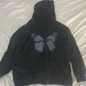 Svart hoodiejacka med blå fjäril - Säljer denna gulliga svarta koftan med en fjäril design på. Det finns ingen lapp så vet inte storleken. Men den är rätt så liten på mig som är runt 165cm, så skulle gissa XXS/XS. Vet inte heller vart den är ifrån. Kontakta gärna innan köp.💗 OBS, katt finns i hemmet!