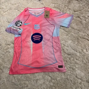 Barça Lamine Yamal tröja rosa/blå Nike M - Snygg FC Barcelona fotbollströja med Lamine Yamal och nummer 10 på ryggen. Tröjan är i rosa och ljusblå toner med Nike-logga, Spotify-sponsor och Champions League-märke på ärmen. Tillverkad i Dri-FIT-material som andas, perfekt för match eller träning.