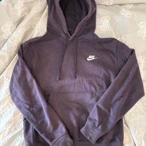 Lila Nike hoodie med broderad logga - Snygg lila hoodie från Nike med broderad vit logga på bröstet. Klassisk känguruficka framtill, huva med snörning och mjukt material som känns skönt mot huden. Har blivit sydd vid magfickan(se bild) men är ej något som syns om man inte, som jag, tar bild med blixt.