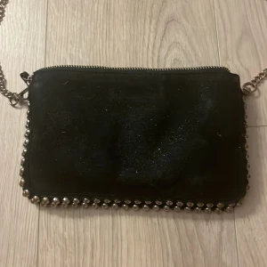 Svart axelväska med kedja och nitar från Zara - Svart axelväska i mocka med silverfärgade nitar längs kanterna och en grov kedja som axelrem från zara. Väskan har dragkedja upptill och flera fack inuti för bra organisering. Perfekt för dig som gillar edgy detaljer och stilren design. Kedjan är ganska rostad men går att byta ut annars är skicket hyfsat bra. Använde ofta förut💞
