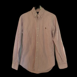 Ralph Lauren skjorta  - Klassisk skjorta från Ralph Lauren i rosa och vitt med smala ränder. Skjortan har lång ärm, knappar hela vägen och broderad logga på bröstet. Tillverkad i bomull och har en normal passform. Perfekt till jeans eller chinos. Jag skickar ej bild när jag bär plaggen och jag vet ej vilken längd och vikt de passar då jag inte prövar kläderna jag säljer, så för storlek hänvisar jag till måtten och passform att man googlar på modellnamn etc 😃