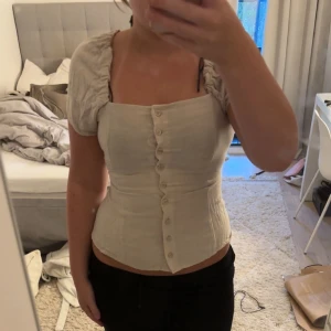 Beige korsettopp med puffärm och knappar - Supersnygg beige korsettopp med puffiga korta ärmar och raka linjer. Toppen har knappar hela vägen framtill och markerad midja för en riktigt smickrande siluett. Perfekt att styla med jeans eller kjol för en trendig look.