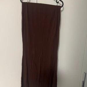 Brun lång kjol - Säljer en snygg brun lång kjol med enkel och stilren design. Klänningen har en lätt rynkad detalj vid bysten och är gjord i ett mjukt, flowy tyg som ger en avslappnad känsla. Perfekt för dig som gillar minimalistisk stil. Storlek L. 