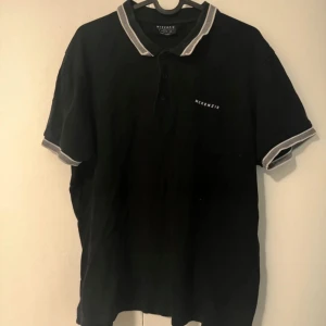 Svart McKenzie pikétröja - Svart piké från McKenzie med vit logga på bröstet. Klassisk krage och knäppning framtill, samt vita detaljer på krage och ärmslut. Perfekt för en clean och sportig stil. Storlek L. 