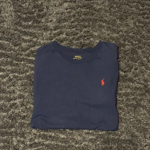 Marinblå t-shirt från Polo Ralph Lauren - Snygg marinblå t-shirt från Polo Ralph Lauren med klassisk röd broderad logga på bröstet. Gjord i mjuk bomull och har en enkel, stilren design med rund hals. Perfekt för dig som gillar clean och tidlös stil.