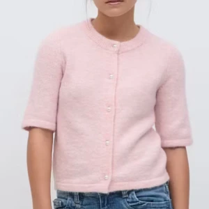 Ljusrosa kofta med pärlknappar - Supersöt ljusrosa kofta med pärlknappar och korta ärmar. Koftan är stickad och har en mjuk, fluffig känsla. Perfekt att slänga över en topp för en gullig look. Rund halsringning och rak passform.