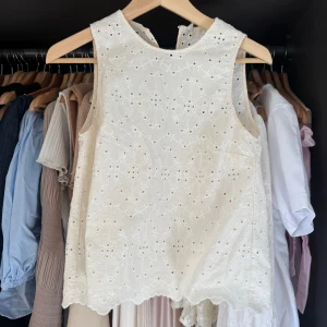 Vit broderad ärmlös blus i bomull - Superfin vit ärmlös blus i bomull med broderade detaljer och hålmönster. Toppen har rund halsringning fram och en liten v-ringning bak. Perfekt för varma dagar och ger en romantisk vibe med sina broderier och vågig nederkant.