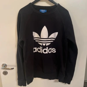 Svart Adidas sweatshirt med logga - Svart sweatshirt från Adidas med stor vit Trefoil-logga framtill. Klassisk rund halsringning och långa ärmar. Tillverkad i mjuk bomullsmix som är skön att ha på sig. Perfekt för en sportig och avslappnad stil.