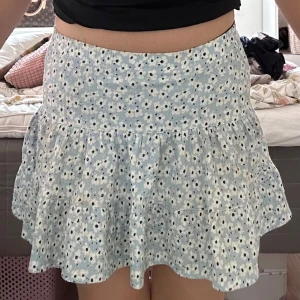 Blommig volangkjol från 157 New - Supersöt kort kjol från 157 New med ljusblå botten och vita blommor med svarta prickar. Kjolen har volanger och smock i midjan för en bekväm passform. Perfekt för dig som gillar en söt och somrig stil!