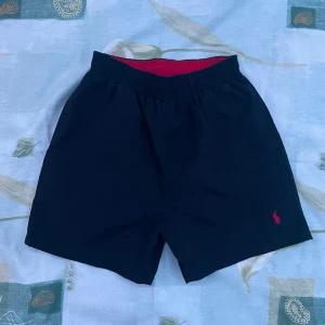Svarta badshorts Polo Ralph Lauren - Snygga Svarta badshorts från Polo Ralph Lauren med röd broderad logga på benet. Elastisk midja med röd insida och meshfoder för extra komfort. Klassisk och stilren design, perfekt för stranden eller poolen.