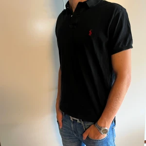 Polo Ralph Lauren piké - Svart t-piké från Polo Ralph Lauren i slim fit-modell. Klassisk pikékrage med två knappar och röd broderad logga på bröstet. Tillverkad i mjuk bomull och har korta ärmar. Perfekt för en clean och stilren look. Pikén har storlek L men modellen har M i storlek.