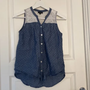 Blå ärmlös blus med spets från KappAhl - Supersöt blå ärmlös blus från KappAhl med vita spetsdetaljer upptill och knappar framtill. Blusen har en lätt A-linjeform och rundad nederkant. Perfekt för dig som gillar romantiska detaljer och vill ha en luftig topp till sommaren.