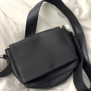 Svart axelväska från C.W. - Stilren svart axelväska från C.W. med bred axelrem och minimalistisk design. Väskan är tillverkad i slätt skinn och har ett lock framtill med diskret logga. Perfekt storlek för det viktigaste och passar till många outfits.