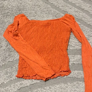 Orange långärmad smocktopp - Trendig orange topp med smockad struktur och långa ärmar. Toppen har en lätt croppad passform och bred, rak urringning. Perfekt för dig som vill sticka ut med färg och cool textur.
