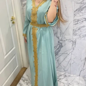 Ljusblå marockansk långklänning med guldiga detaljer - Säljer en elegant ljusblå marockansk långklänning med vackra guldiga broderier längs v-ringningen, ärmsluten och framtill. Klänningen har ett matchande bälte i midjan och långa ärmar. Perfekt för dig som vill sticka ut med en  lyxig look.✨