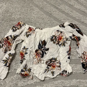Offshoulder blommig topp i vitt - Superfin offshoulder topp i vitt med stora blommiga mönster i rött, beige och mörkblått. Långa ärmar och smockad resår vid axlar och midja ger en snygg passform. Perfekt för dig som gillar romantisk och trendig stil.