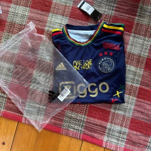 Ajax x Bob Marley svart fotbollströja - Unik svart Ajax fotbollströja från Adidas med Bob Marley-tema. Färgglada ränder i rött, gult och grönt på axlar och ärmslut, tryck med texter och illustrationer, samt Ajax-logga och Ziggo-sponsor. Tillverkad i lätt funktionsmaterial.