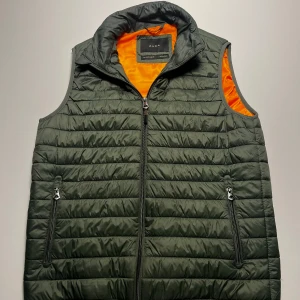 Grön dunväst från Zara med orange foder - Snygg quiltad dunväst från Zara i mörkgrönt med kontrasterande orange foder. Västen har dragkedja framtill, två fickor med dragkedja och hög krage. Perfekt för lager-på-lager och ger en cool streetstyle-vibe.