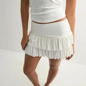 Vit volangkjol  - Supersöt vit kortkjol med dubbla volanger och bred midja. Kjolen är gjord i mjuk bomull och har en flowy känsla som ger en chill och trendig look. Perfekt att styla med crop top eller linne för en fräsch vibe.