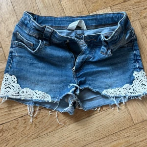 Jeansshorts med spetsdetaljer - Blå jeansshorts med råa kanter och snygga vita spetsdetaljer på benen. Klassisk femficksmodell med dragkedja och bälteshällor. Perfekta för sommaren och ger en chill vibe med den slitna looken och de dekorativa spetsinslagen.