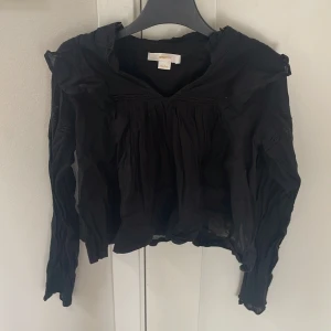 Svart blus med volang från Gina Tricot - Svart blus från Gina Tricot med långa ärmar, volangdetaljer vid axlarna och spetsinslag på både ärmar och över bröstet. Blusen har en luftig och lätt rynkad design som ger en cool och avslappnad vibe.