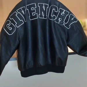Givenchy svart skinn bomberjacka - Exklusiv svart bomberjacka från Givenchy i äkta skinn med stora vita bokstäver på ryggen. Jackan har tryckknappar framtill, ribbade muddar och klassisk bomberkrage. Perfekt statement-plagg för dig som vill sticka ut med lyxig streetstyle.