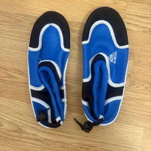 Blå badskor från Billabong - Blå badskor från Billabong med svarta och vita detaljer. Skorna har vit räfflad sula och justerbar dragsko bak för perfekt passform. Perfekta för stranden eller poolen, tillverkade i syntetmaterial som torkar snabbt.