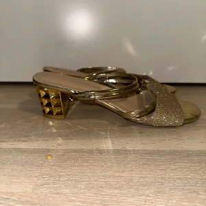 Glittriga guldiga sandaler med klack - Snygga sandaler i guld med glittrig bred rem över tårna och två smala metallicband. Klacken är låg och dekorerad med pyramidformade detaljer för extra edge. Öppen tå och platt sula framtill, perfekta för att sticka ut.