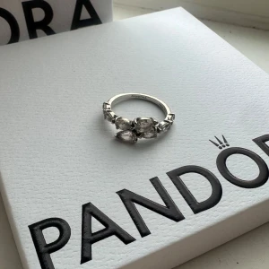 Pandora silverring med stenar - Superfin silverring från Pandora med flera stenar i en snygg blomliknande design. Kommer i originalask från Pandora och även med påse ifall man skulle vilja ge bort den. Den är i väldigt fint skick och använd med omsorg. Är inte helt hundra på storleken men tror att det var 17mm om jag minns rätt. 