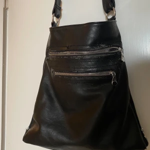 Svart axelväska i skinn med dragkedjor - Stilren svart axelväska i skinn med flera silvriga dragkedjor framtill och baktill. Väskan har en mjuk, lite slouchy form och bred axelrem med metallringar. Perfekt för dig som vill ha plats för allt du behöver och gillar en clean look. Frakt tillkommer 