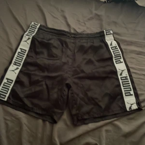 Svarta shorts från Puma med logoband - Svarta shorts från Puma med breda vita logoband längs sidorna där det står PUMA och har loggan. Klassisk sportig look, elastisk midja och ledig passform. Perfekta för träning eller chill.