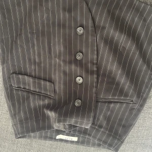 Svart randig kavaj från NA-KD - Snygg svart kavaj från NA-KD med tunna vita ränder. Klassisk passform med fyra knappar framtill och två fickor med lock. Perfekt för dig som gillar en stilren och trendig look.
