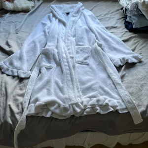 Vit morgonrock med volang och huva - Mysig vit morgonrock i frotté med volangkant längs ärmar och nederkant samt knytbälte i midjan. Perfekt för chill dagar hemma eller efter duschen. Har två fickor framtill och en skön, avslappnad passform.