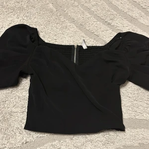 Svart croppad blus med puffärmar - Säljer en svart croppad blus med puffiga korta ärmar och smockad rygg. Toppen har en snygg omlott framtill och dragkedja med ringdetalj bak. Perfekt för dig som gillar trendiga detaljer och en feminin siluett.