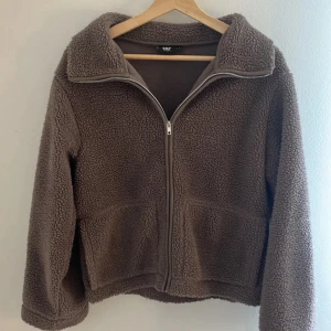Brun teddyjacka från 157, storlek S - !!INTE MIN BILD!! Mysig brun teddyjacka från 157 i storlek S. Jackan har dragkedja framtill, två stora fickor och en hög krage. Perfekt för kyliga dagar när du vill vara både varm och snygg. Den fluffiga teddytexturen ger en chill och trendig vibe.