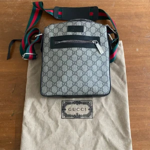Gucci axelväska med GG-mönster - Snygg axelväska från Gucci med klassiskt GG-mönster i grått och svart. Väskan har en dragkedja framtill och justerbar axelrem i grönt och rött. Perfekt för dig som vill ha en stilren och ikonisk accessoar.