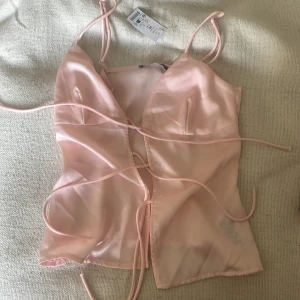 Ljusrosa satinlinnetopp med snörning - Supersöt ljusrosa linne-topp i satin från gina med tunna axelband och trendig snörning framtill. Toppen har v-ringning och croppad passform, perfekt att styla med jeans eller kjol för en cool look. Sjukt söt och oanvänd 