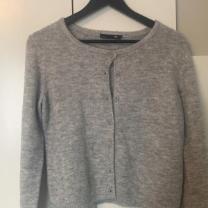 Ljusgrå kofta från 157, XS - Mysig ljusgrå kofta från 157 i stickad ullmix. Klassisk rund halsringning, knäppning framtill med enkla knappar och långa ärmar. Perfekt att slänga över en t-shirt eller linne för extra värme under kyliga dagar.