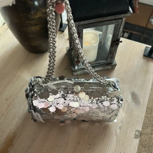 Silver paljettväska från BikBok - Glittrig axelväska från BikBok i silver med stora och små paljetter över hela väskan. Kedjehandtag i metall och cylinderformad design. Perfekt för att sticka ut och ge din outfit extra bling. Stängs med dragkedja och har ett rymligt fack. HELT NY!!