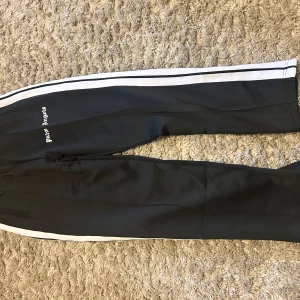 Palm Angels svarta trackpants med ränder - Svarta trackpants från Palm Angels med vita sidoränder och logga tryckt på låret. Byxorna har elastisk midja och är gjorda i ett glansigt syntetmaterial. Perfekta för en sportig och trendig streetwear-look.