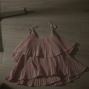 Ljusrosa plisserad linnetopp - Supersöt ljusrosa linnetopp med plisserade volanger och tunna axelband som knyts upptill. Toppen har en flowy och luftig känsla, perfekt för varma dagar. Den har en romantisk vibe med sina lager och mjuka pastellton.