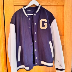 Blåvit bomberjacka med brodyr - Snygg blå bomberjacka med vita ärmar och broderad G på bröstet. Baksidan har cool brodyr med texten 'Newington Georgia'. Jackan har ribbade muddar, tryckknappar och kontrastfärgade detaljer. Perfekt för dig som gillar college-stil. Lite smutts på armen 